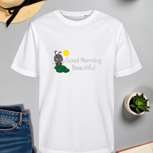 Bijela majica s natpisom "Good Morning Beautiful" u sivom fontu, popraćena ilustracijom simpatičnog crnog kukca s crvenim bubamarastim krilima, koji sjedi na zelenom listu dok sunce sja iznad njega. Majica je položena na ravnu površinu uz slameni šešir s crnom vrpcom, svijeću, zelenu biljku u tegli, otvorenu knjigu i traperice.