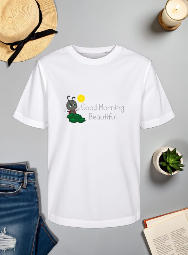 Bijela majica s natpisom "Good Morning Beautiful" u sivom fontu, popraćena ilustracijom simpatičnog crnog kukca s crvenim bubamarastim krilima, koji sjedi na zelenom listu dok sunce sja iznad njega. Majica je položena na ravnu površinu uz slameni šešir s crnom vrpcom, svijeću, zelenu biljku u tegli, otvorenu knjigu i traperice.