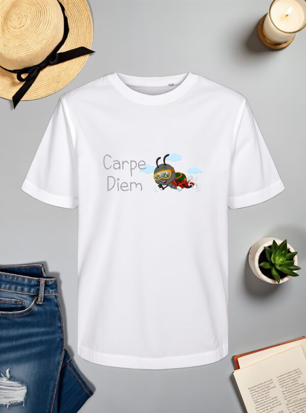 Bijela majica s natpisom "Carpe Diem" u sivom fontu, popraćena ilustracijom simpatičnog crnog kukca s crvenim bubamarastim krilima, koji nosi padobransku opremu i zaštitne naočale dok leti kroz zrak s oblacima u pozadini. Majica je položena na ravnu površinu uz slameni šešir s crnom vrpcom, svijeću, zelenu biljku u tegli, otvorenu knjigu i traperice.