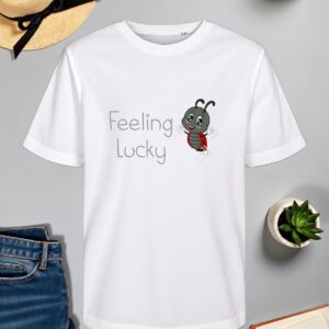 Bijela majica s natpisom "Feeling Lucky" u sivom fontu, popraćena ilustracijom vesele bubamare s crvenim krilima i crnim točkicama koja skače s osmijehom na licu. Majica je položena na ravnu površinu uz slameni šešir s crnom vrpcom, svijeću, zelenu biljku u tegli, otvorenu knjigu i traperice.