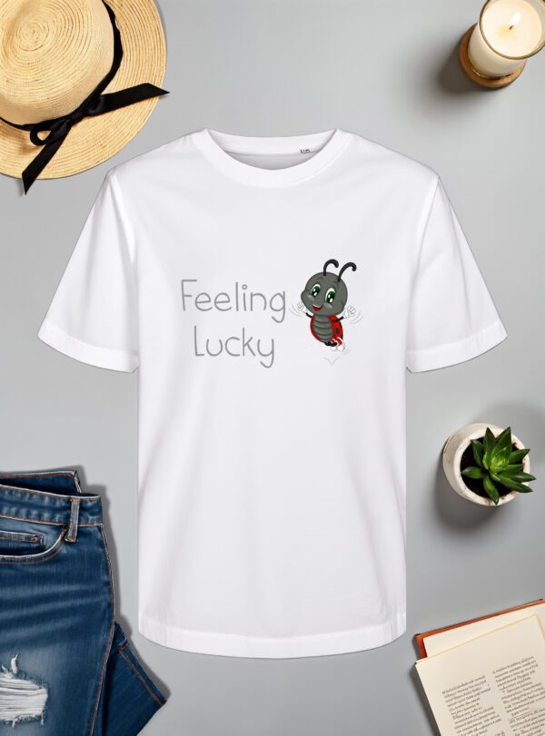 Bijela majica s natpisom "Feeling Lucky" u sivom fontu, popraćena ilustracijom vesele bubamare s crvenim krilima i crnim točkicama koja skače s osmijehom na licu. Majica je položena na ravnu površinu uz slameni šešir s crnom vrpcom, svijeću, zelenu biljku u tegli, otvorenu knjigu i traperice.