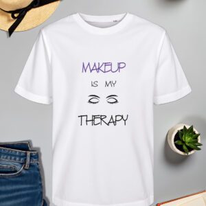 Bijela majica s natpisom "MAKEUP IS MY THERAPY" u kombinaciji ljubičaste i crne boje. Dizajn uključuje stilizirane ilustracije zatvorenih očiju s dugim trepavicama, naglašavajući ljubav prema šminkanju. Majica je položena na ravnu površinu uz slameni šešir s crnom vrpcom, svijeću, zelenu biljku u tegli, otvorenu knjigu i traperice.