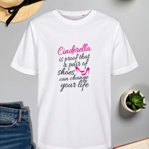 Bijela majica s natpisom *"Cinderella is proof that a pair of shoes can change your life"* ispisanim elegantnim crnim i ružičastim fontom. U dizajnu je uključena stilizirana ilustracija ružičaste cipele, naglašavajući motiv inspiriran Pepeljugom. Majica je položena na ravnu površinu uz slameni šešir s crnom vrpcom, svijeću, zelenu biljku u tegli, otvorenu knjigu i traperice.