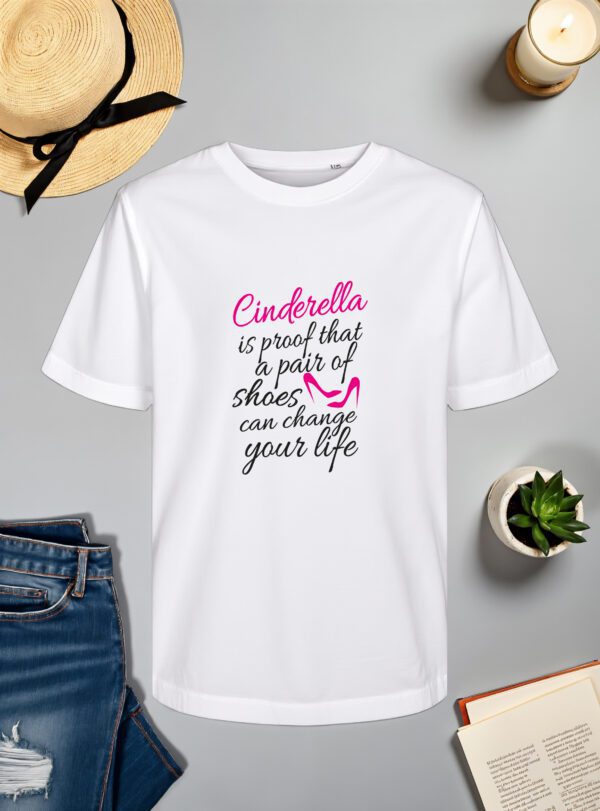 Bijela majica s natpisom *"Cinderella is proof that a pair of shoes can change your life"* ispisanim elegantnim crnim i ružičastim fontom. U dizajnu je uključena stilizirana ilustracija ružičaste cipele, naglašavajući motiv inspiriran Pepeljugom. Majica je položena na ravnu površinu uz slameni šešir s crnom vrpcom, svijeću, zelenu biljku u tegli, otvorenu knjigu i traperice.