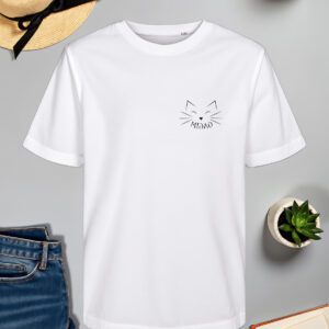 Bijela majica s minimalističkim crnim printom mačjeg lica smještenim na lijevoj strani prsa. Dizajn uključuje jednostavne linije koje oblikuju mačje oči, brkove i nos, uz natpis "MIAU" ispod. Majica je položena na ravnu površinu uz slameni šešir s crnom vrpcom, svijeću, zelenu biljku u tegli, otvorenu knjigu i traperice.