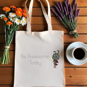 Platnena vrećica s natpisom *"Be Awesome Today"*, ilustracijom simpatične bubamare koja drži malo ogledalo i nosi ruksak. Vrećica je položena na drveni stol, okružena buketima tratinčica, narančastih cvjetova i lavande, uz šalicu crne kave na bijelom tanjuriću.