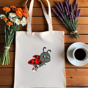 Platnena vrećica s ilustracijom simpatične bubamare u crveno-crnim krilima i tenisicama, koja leti i veselo maše. Vrećica je položena na drveni stol, okružena buketima tratinčica, narančastih cvjetova i lavande, uz šalicu crne kave na bijelom tanjuriću.