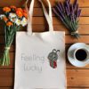 Platnena vrećica s natpisom *"Feeling Lucky"*, ilustracijom simpatične bubamare u crveno-crnom kaputiću koja veselo skače. Vrećica je položena na drveni stol, okružena buketima tratinčica, narančastih cvjetova i lavande, uz šalicu crne kave na bijelom tanjuriću.