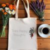 Platnena vrećica s motivacijskim natpisom *"Think Happy Thoughts"* i ilustracijom simpatične bubamare u crveno-crnim krilima i tenisicama, zamišljene i nasmijane. Vrećica je položena na drveni stol, okružena buketima tratinčica, narančastih cvjetova i lavande, uz šalicu crne kave na bijelom tanjuriću.