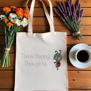 Platnena vrećica s motivacijskim natpisom *"Think Happy Thoughts"* i ilustracijom simpatične bubamare u crveno-crnim krilima i tenisicama, zamišljene i nasmijane. Vrećica je položena na drveni stol, okružena buketima tratinčica, narančastih cvjetova i lavande, uz šalicu crne kave na bijelom tanjuriću.