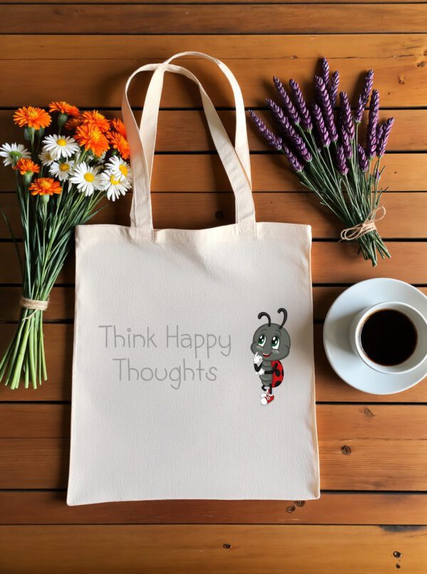 Platnena vrećica s motivacijskim natpisom *"Think Happy Thoughts"* i ilustracijom simpatične bubamare u crveno-crnim krilima i tenisicama, zamišljene i nasmijane. Vrećica je položena na drveni stol, okružena buketima tratinčica, narančastih cvjetova i lavande, uz šalicu crne kave na bijelom tanjuriću.