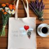 Platnena vrećica s natpisom "Love is in the Air" i ilustracijom simpatične bubamare koja drži crveni balon u obliku srca, okružena oblacima. Vrećica je položena na drveni stol, uz bukete tratinčica, narančastih cvjetova i lavande, te šalicu crne kave na bijelom tanjuriću.