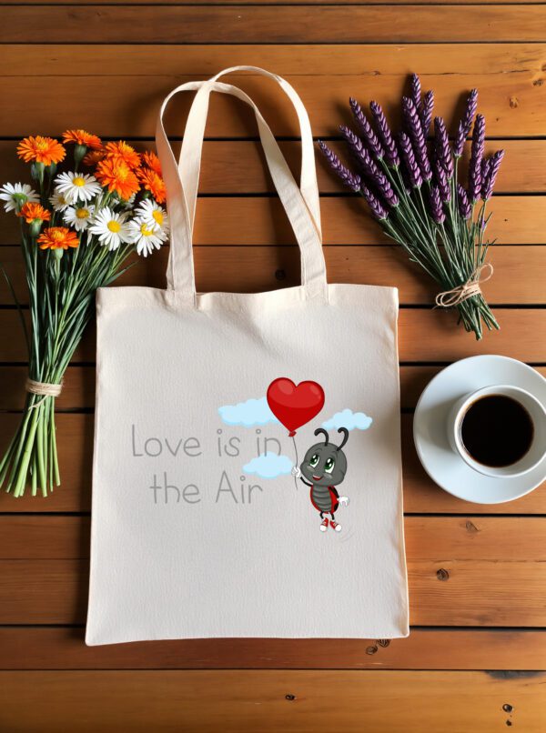 Platnena vrećica s natpisom "Love is in the Air" i ilustracijom simpatične bubamare koja drži crveni balon u obliku srca, okružena oblacima. Vrećica je položena na drveni stol, uz bukete tratinčica, narančastih cvjetova i lavande, te šalicu crne kave na bijelom tanjuriću.