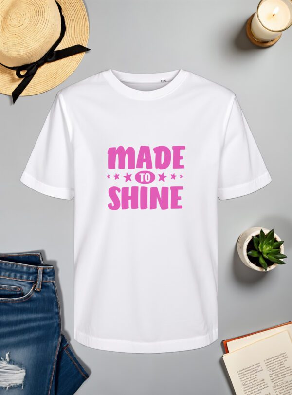 bijela majica s natpisom "MADE TO SHINE" u ružičastoj boji, ukrašenom zvjezdicama oko riječi "TO". Majica je položena na ravnu površinu uz slameni šešir s crnom vrpcom, svijeću, zelenu biljku u tegli, otvorenu knjigu i traperice.