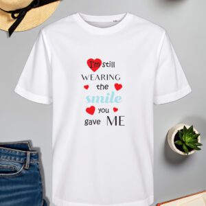 Bijela majica s natpisom I'm still wearing the smile you gave me, ukrašena crvenim srcima i plavim slovima, položena na ravnu površinu uz modne dodatke.