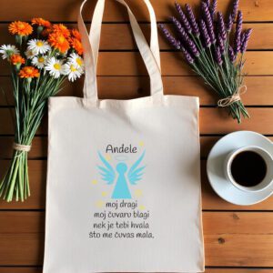 Bež platnena vrećica s tiskanim dizajnom leži na drvenoj podlozi. Na vrećici je stilizirani plavi anđeo s aureolom iznad glave i raširenim krilima, okružen žutim zvjezdicama. Iznad anđela ispisana je riječ 'Anđele' crnim slovima, a ispod se nalazi tekst: moj dragi moj čuvaru blagi nek je tebi hvala što me čuvaš mala.