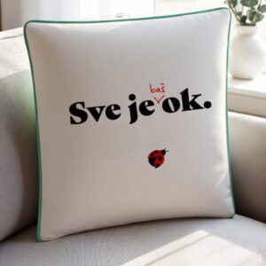 Dekorativni jastuk s natpisom "Sve je baš ok." i ilustracijom bubamare na bijeloj pozadini, postavljen na svijetloj sofi. Stražnja strana jastuka je u *sage green* nijansi, dodajući umirujući ton prostoru.