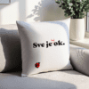 Bijeli dekorativni jastuk s natpisom "Sve je baš ok." u crno-crvenoj boji i ilustracijom bubamare, smješten na svijetloj sofi uz prozor s biljkama u pozadini.