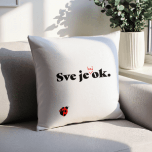 Bijeli dekorativni jastuk s natpisom "Sve je baš ok." u crno-crvenoj boji i ilustracijom bubamare, smješten na svijetloj sofi uz prozor s biljkama u pozadini.