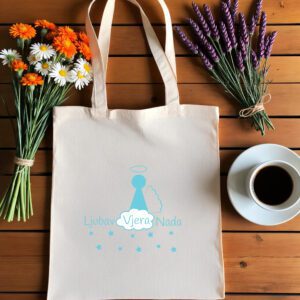 Bež platnena vrećica s tiskanim dizajnom leži na drvenoj podlozi. Na vrećici je stilizirani plavi anđeo s aureolom iznad glave, s jednim krilom, okružen zvjezdicama. Ispod anđela nalazi se bijeli oblak s istaknutom riječju 'Vjera', dok su riječi 'Ljubav' i 'Nada' napisane svijetloplavom bojom s obje strane oblaka.