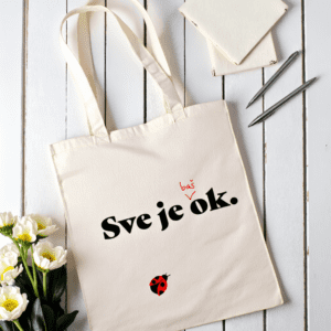 Platnena torba u bež boji s natpisom Sve je baš ok. i ilustracijom bubamare, položena na bijelom drvenom stolu uz bilježnice, olovke i bijele cvjetove.