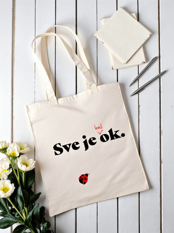 Platnena torba u bež boji s natpisom Sve je baš ok. i ilustracijom bubamare, položena na bijelom drvenom stolu uz bilježnice, olovke i bijele cvjetove.