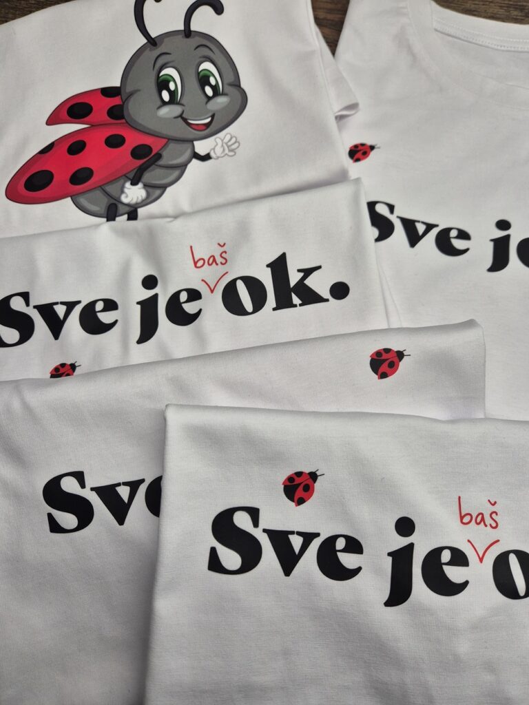 Bijele majice s tiskom natpisa "Sve je baš OK" i motivom bubamare raspoređene na drvenoj površini. Jedna majica sadrži ilustraciju simpatične bubamare s osmijehom i podignutom rukom. Dizajn kombinira crnu i crvenu boju, stvarajući upečatljiv vizualni identitet.