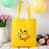 žuta platnena vrećica s motivom veselog žutog pileta koje drži bijelu tratinčicu. Vrećica je postavljena na drveni stol, okružena pastelno obojenim balonima i cvijećem u pozadini.