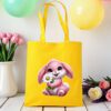 žuta platnena vrećica s motivom preslatkog rozog zeca koji drži bijelu tratinčicu. Vrećica je postavljena na drveni stol, okružena pastelno obojenim balonima i cvijećem u pozadini.