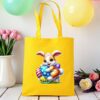 žuta platnena vrećica s uskršnjim motivom – razigrani smeđi zeko s plavim očima sjedi na travi, okružen šarenim pisanicama. Vrećica je postavljena na drveni stol, uz pastelno obojene balone i cvjetni aranžman u pozadini.