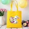 žuta platnena vrećica s uskršnjim motivom – simpatični bijeli zeko grli veliku, šareno ukrašenu pisanicu. Vrećica je postavljena na drveni stol, okružena pastelno obojenim balonima i cvijećem u pozadini.